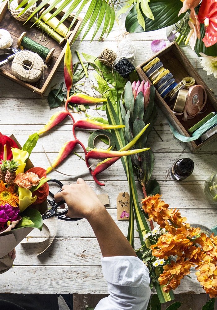 A hand arranges a vibrant flower bouquet on a rustic wooden table filled with various tropical flowers, greenery, twine, ribbons, scissors, and a small "thank you" tag.