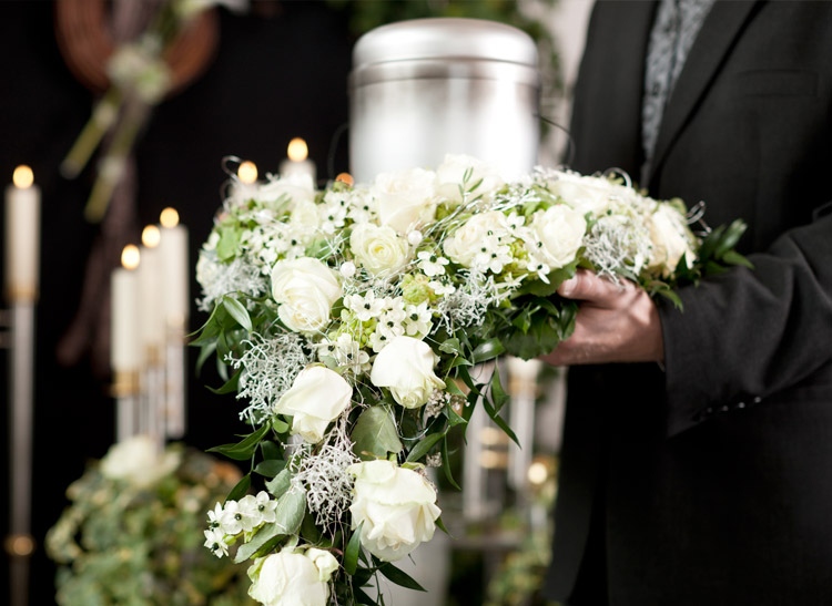 A person in a black suit holds a white floral wreath in front of a silver urn, with lit candles in the blurred background.