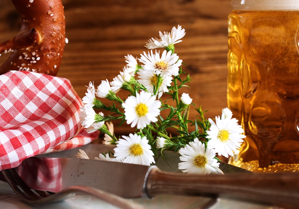 A soft pretzel and checkered napkin rest on a wooden table beside white daisies, a knife, and a glass mug filled with foamy beer in a rustic setting.