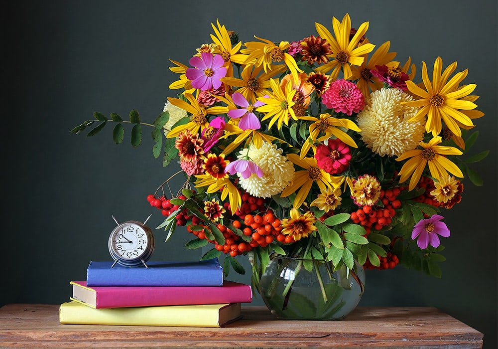 A colorful bouquet of various flowers in a glass vase, placed on a wooden table, alongside a stack of three books and an analog clock.