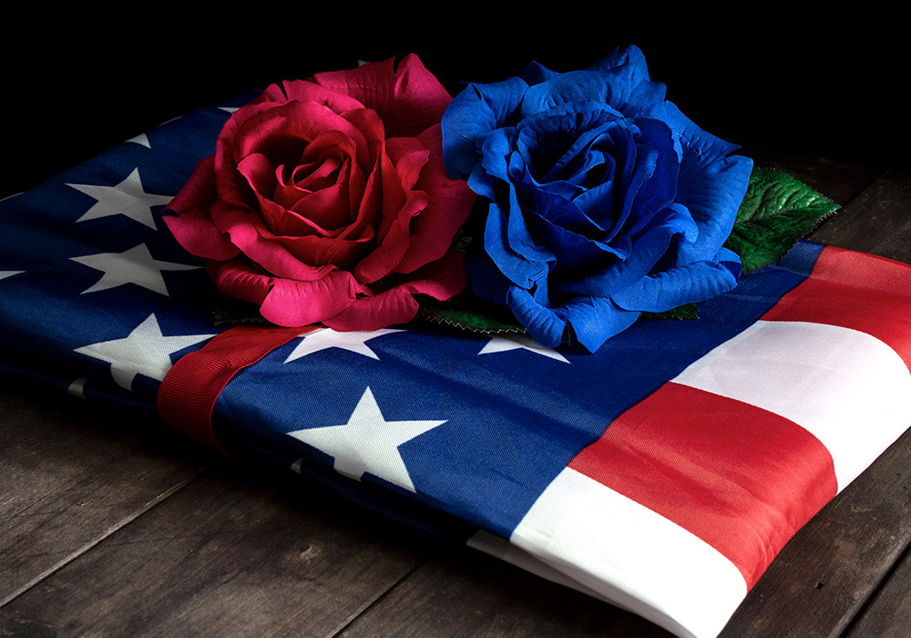 Two large artificial roses, one red and one blue, rest atop a folded American flag with white stars, situated on a wooden surface.