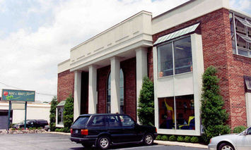 A brick building with tall white columns has a black car parked in front. The building is surrounded by greenery and another building with a sign that reads 