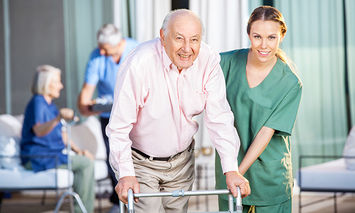 An elderly man using a walker is assisted by a smiling nurse in green scrubs. In the background, another elderly woman and caregiver are seated.