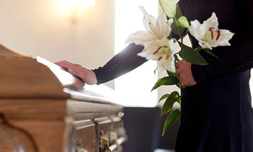 A person in black attire holds white lilies while touching a wooden casket in a well-lit room, likely a funeral setting.